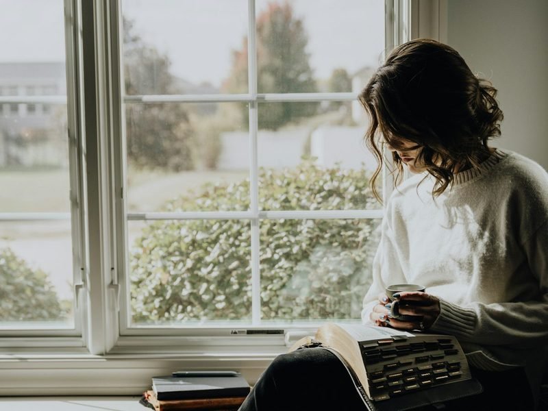 A woman sitting by a large window with a book and a warm drink
