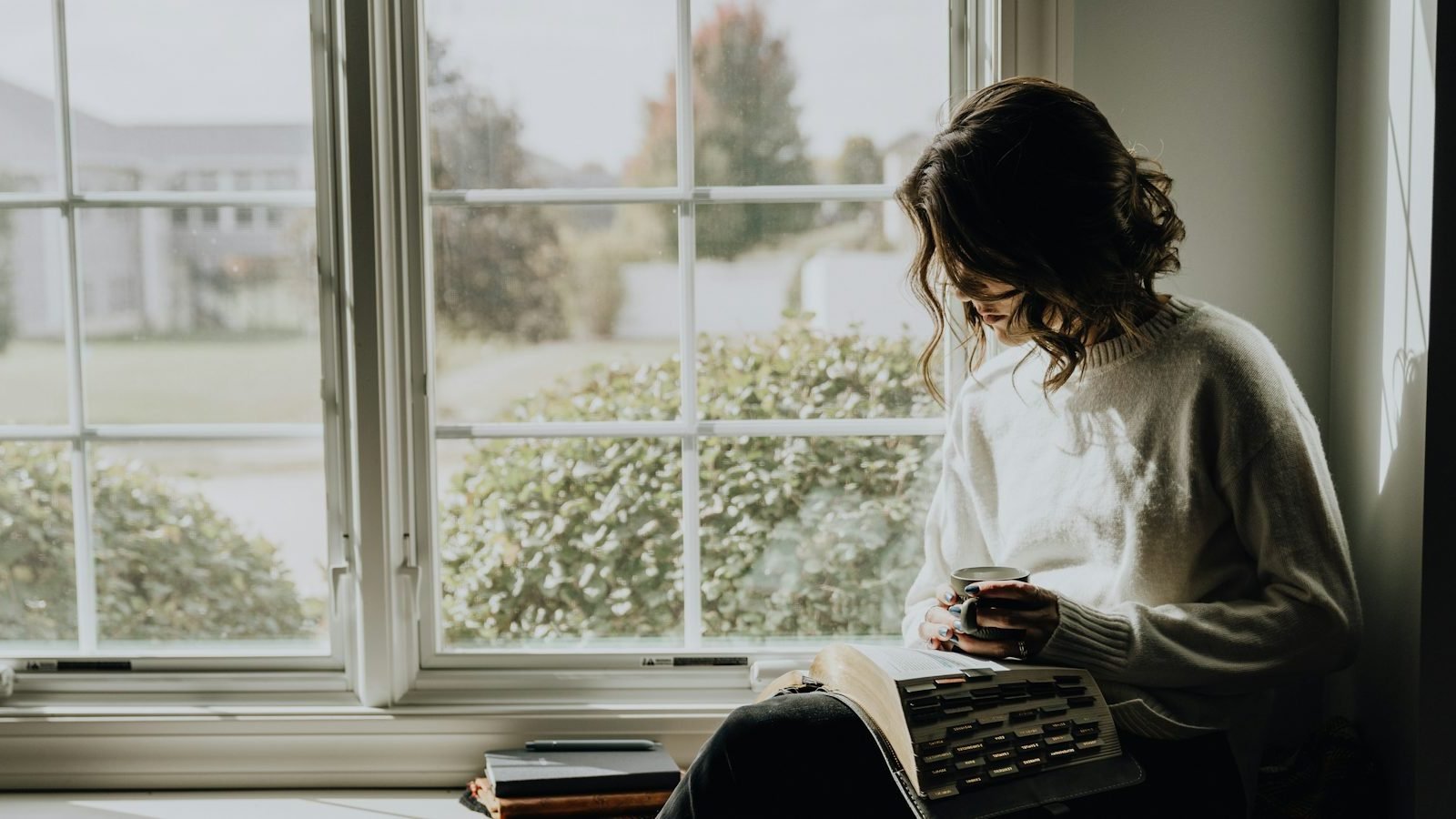 A woman sitting by a large window with a book and a warm drink