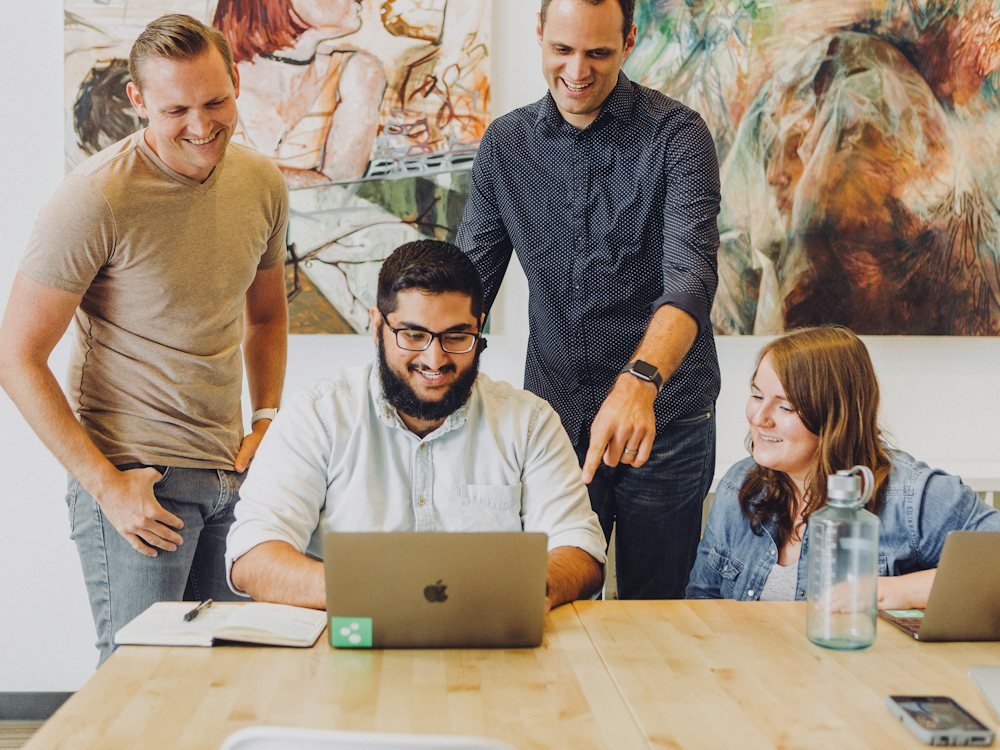 Four colleagues gathered around a laptop, smiling and talking through an idea