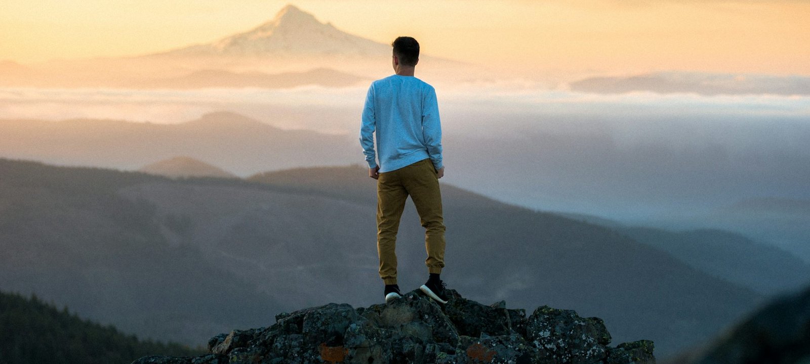A man standing on a rocky outcrop, looking out over the hills at sunrise