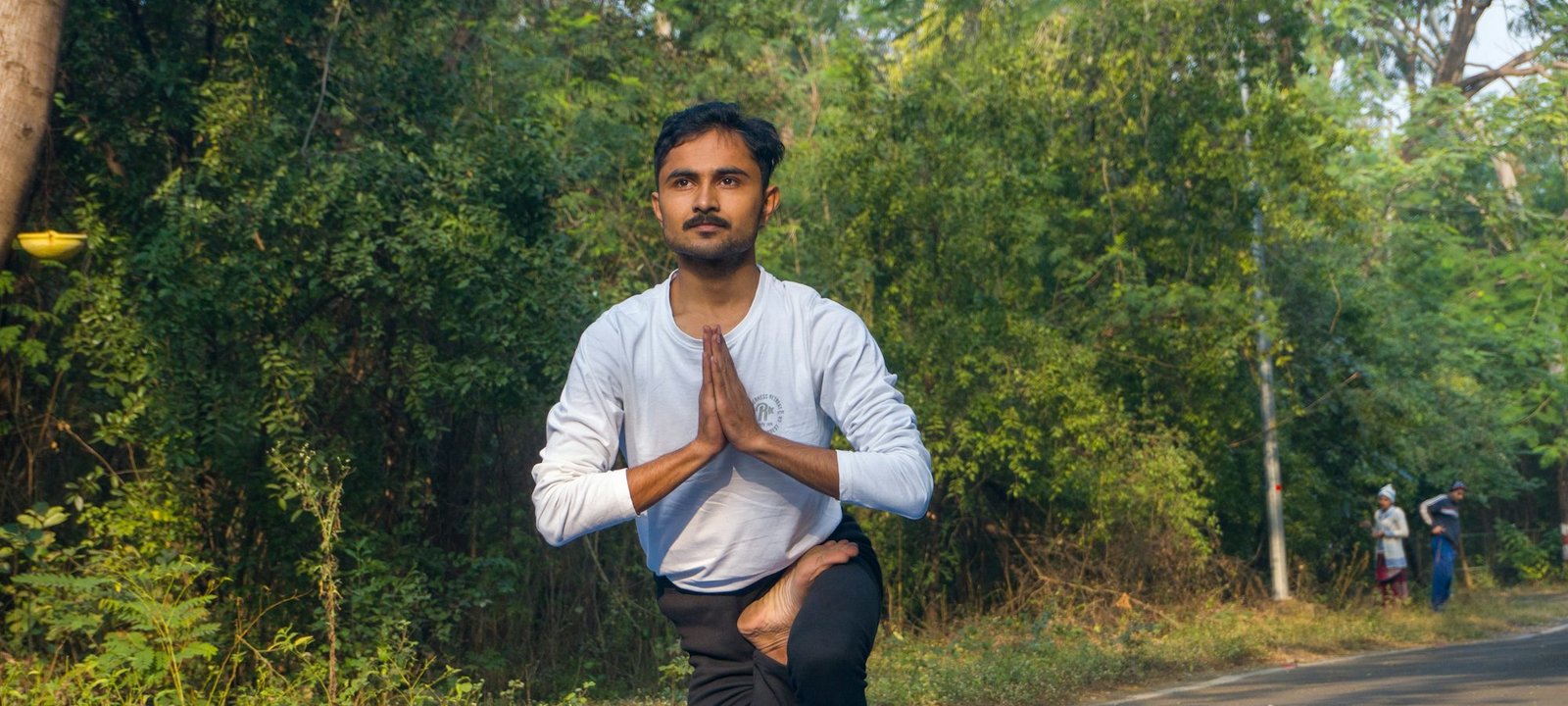 A young man kneeling on a yoga mat outdoors, hands together, eyes calm