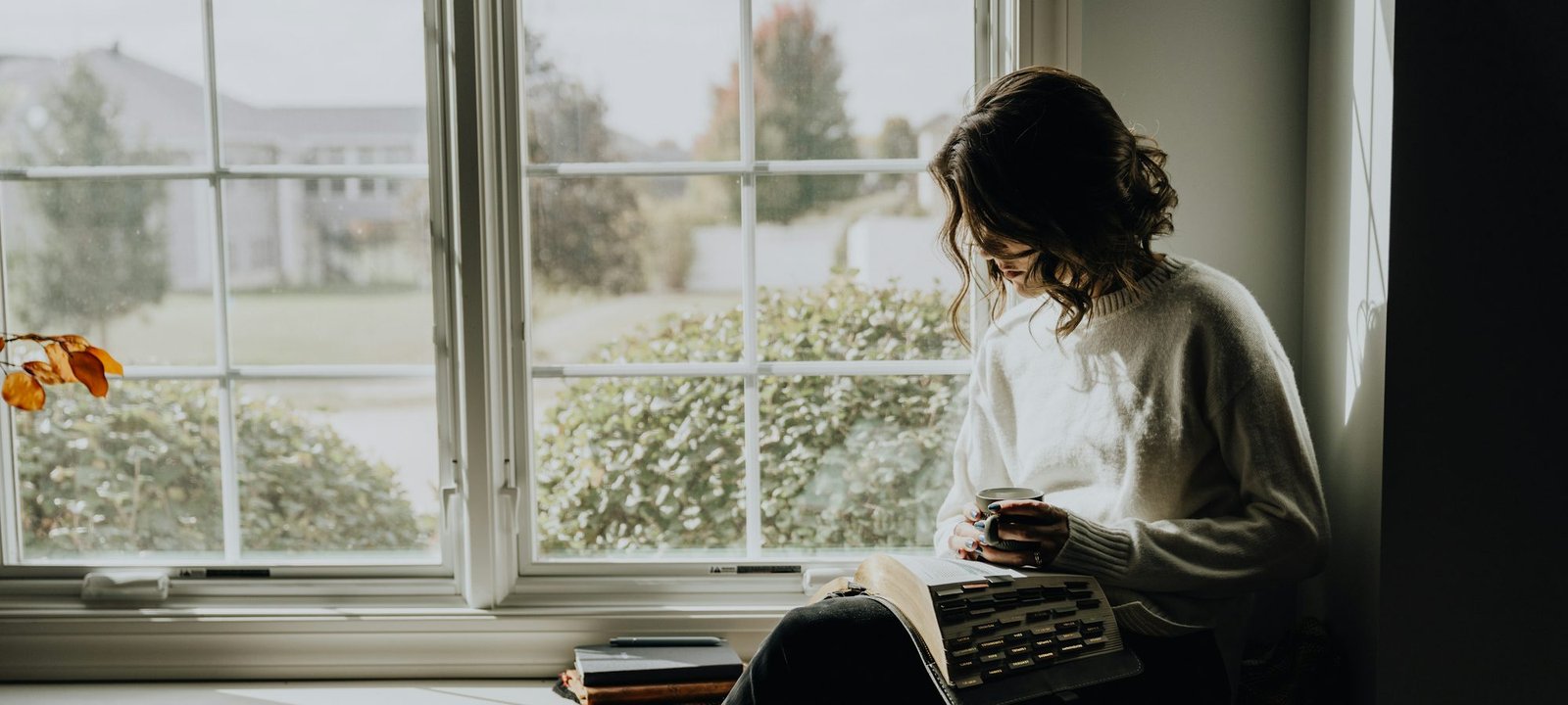 A woman sitting by a large window with a book and a warm drink