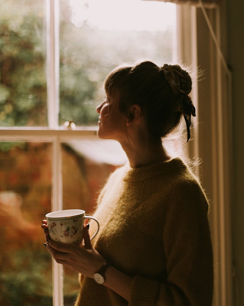 A woman holding a cup of tea, reflecting by a window